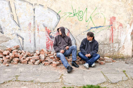 two street hooligans or rappers standing against a graffiti painted wall are preparing to smoke a cigaretteの写真素材