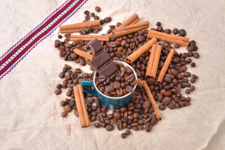 Cup of coffee with cinnamon sticks, bitten bar of chocolate on vintage texture. Roasted coffee beans...