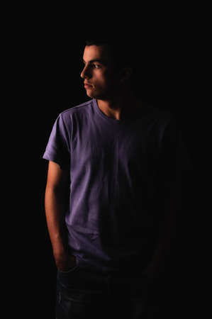 Portrait of young man wearing t-shirt standing with hands in pockets and looking on one side isolated on black backgroundの写真素材