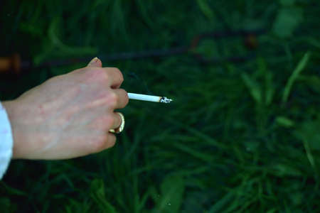 Cigarette in the hand of a woman, enjoying her cigarette in the morning on the green grass background close up. Lights and shadows. Selective focusの写真素材