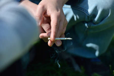 Hand of a man holding a cigarette in hand outside, on a background of green grass. Unhealthy habit. selective focusの写真素材