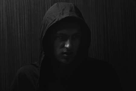 Picture of a teenage boy with serious expression and black hoodie on his head, direct gaze.Black and white. Dark background, lights and shadowsの写真素材