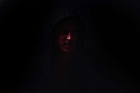 Portrait of teenage boy with serious expression and black hoodie on his head, brown dark hair, direct gaze. Dark background, lights and shadowsの写真素材