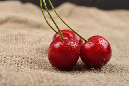 Horizontal photo of tree red cherries with water drops which are together on one green branch on jute bon a table. Selective focus. Lights and shadowsの写真素材