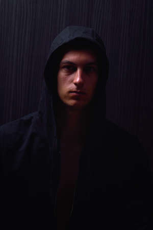 Portrait of teenage boy with serious expression and black hoodie on his head, brown dark hair, direct gaze. Dark background, lights and shadowsの写真素材