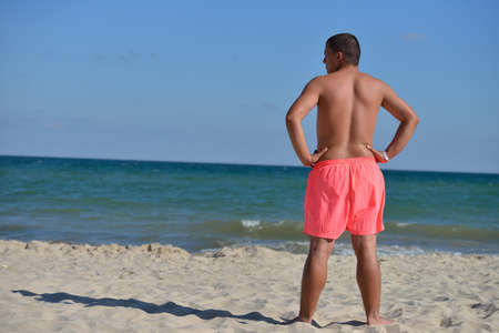 Athletic young man in pink shorts sits on the beach and looks at the sea, he prepares to go and swim. Burning sun, holidayの写真素材