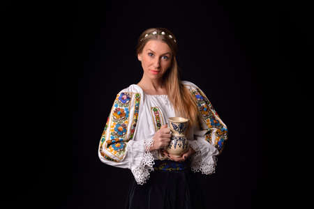 Closeup of a young Romanian woman dressed in traditional costume, holding in his hands a cup of painted clay. Romanian folklore. Posing in a studio on a black backgroundの写真素材