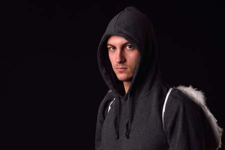 Young man with black hoodie and white feathered wings behind the back on a dark background. Conceptual photo, good and badの写真素材