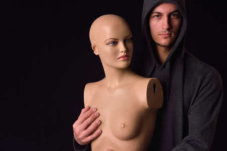Serious man dressed in black hoodie holding in his arms a plastic women body mannequin on dark background. Lonelinessの写真素材
