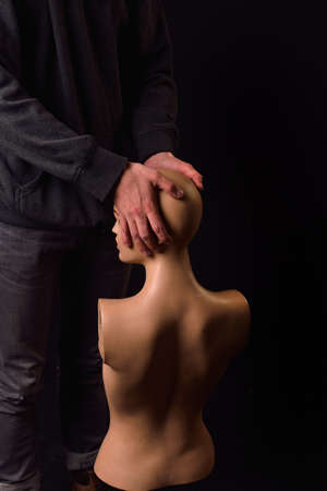 Serious man dressed in black hoodie holding in his arms a plastic women body mannequin on dark background. Lonelinessの写真素材