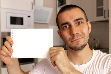 Young man holding and show blank paper notepad with space for text. Kitchen background blurred.