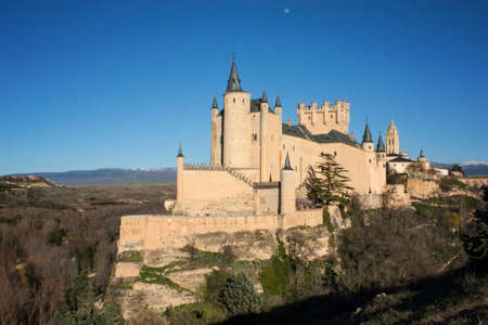 Beautiful medieval stone castle on top of rocks with mountains in the background. A blue sky with the moon and surrounded by forest. Segovia, Spainのeditorial素材