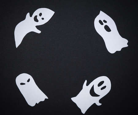 Four White Ghosts On Black Paper Background. Flat Lay Top View Minimal Style. Creative Halloween Concept. Scary, Fun, Smiling And Horror Faces. Handmade Artwork. Copy Spaceの写真素材