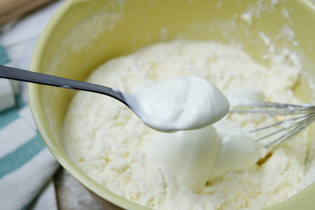 Adding spoon of sour cream to the dough. Process of mixing eggs, preparing pie. Top view with light...