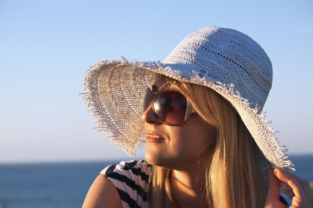Young smiling blond caucasian white woman with hat and sunglasses looking at sunset. Clear blue sky...