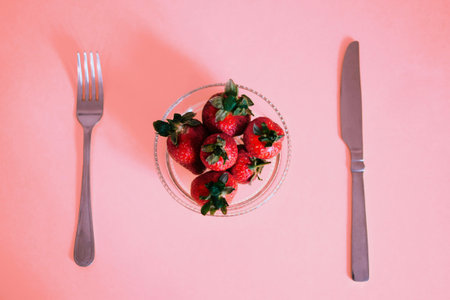 Strawberries in a glass bowl with a knife and fork on a pink background