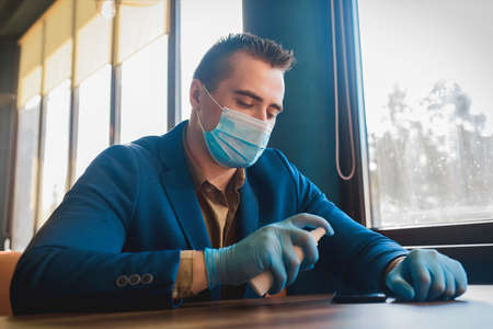 Young european businessman guy in a blue suit, a protective mask and medical gloves holds an antiseptic in his hand and sprays on a mobile phone.の写真素材