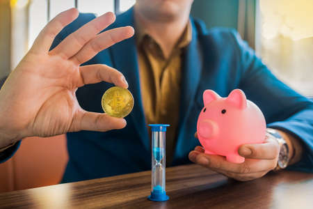 A businessman's hands are holding a gold cryptocurrency bitcoin and a pig piggy bank next to an hourglass on the table. Bitcoin and cryptocurrency concept.の写真素材
