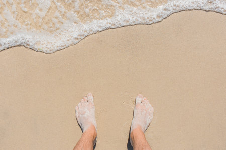 The young man's legs stand on the beach sand and are washed by blue sea water, top view.