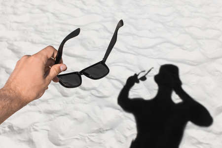 The young man's hand holds sunglasses against the background of his black silhouette shadow on the white beach sand.の写真素材