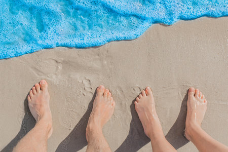 Men's and women's feet stand next to each other on wet beach sand near blue sea water, top view.