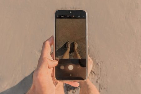 The man's hand holds a mobile phone and takes pictures of his bare feet standing on the wet beach...