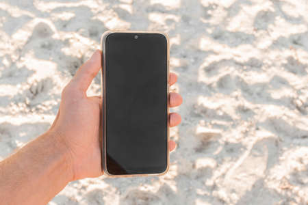 The hand holds a mobile phone with a black screen, a place for text and design, against the background of white beach sand.の写真素材