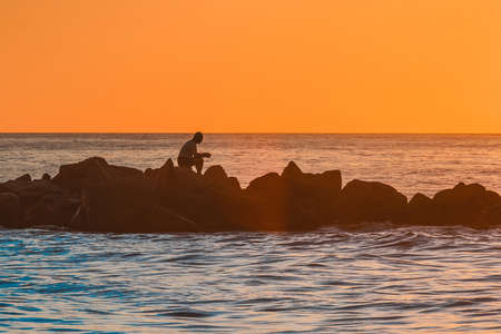 The silhouette of a lonely man sitting in meditation on the rocks of a breakwater near the sea coast against the orange sunset of the sky and horizon line background.の写真素材