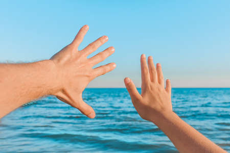 The hand of a guy and a girl stretch to the sea or wave to him against the blue sky background and the horizon line at sunset, close up.の写真素材
