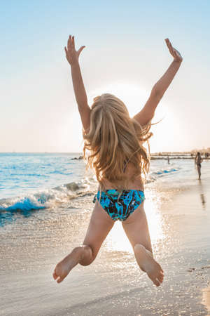 Young happy girl in swimsuit jumps her arms to the sides on the beach against the backdrop of the sea and sunset.の写真素材