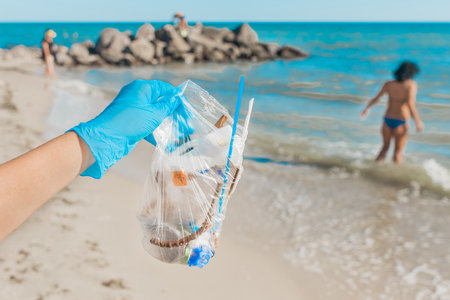 A man's hand in protective latex gloves holds a bag of waste against the sea. Cleaning up the garbage behind people.の写真素材