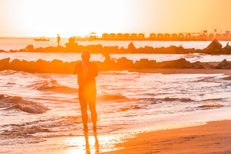 The silhouette of one single man in a cap stands on the seashore by the water and looks at the orange sky sunset.の写真素材