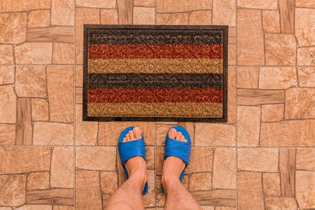 Male feet in blue house slippers stand in front of a foot mat on a brown tiled floor texture background, top view.の写真素材