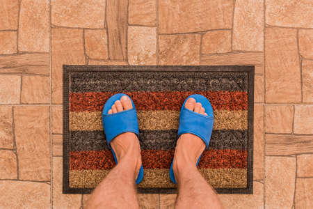 Male feet in blue house slippers stand on a foot mat on a brown tiled floor texture background, top view.の写真素材