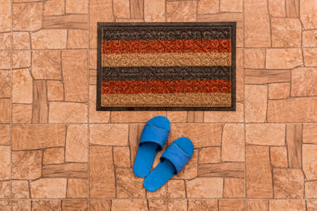 Men's blue house slippers stand near foot mat on a brown tiled floor texture background, top view.の写真素材
