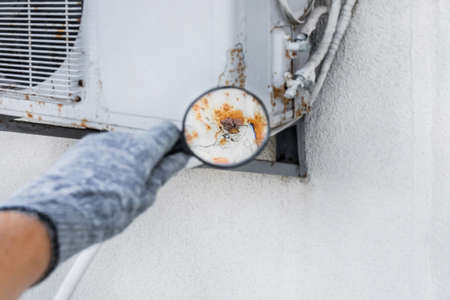 Hand of industrial worker in construction glove with magnifying glass examines rust on old air conditioner against white wall background.の写真素材