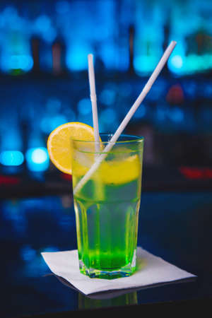 Cooling alcoholic cocktail with green syrup and lemon on a bar counter against a blue nightclub background.の写真素材