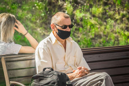 Belarus, Minsk region - July 18, 2020: An adult man in a black protective mask and dark glasses sits on a bench with folded arms.のeditorial素材