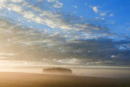 Dawn in a field with the light of the sun from beyond the horizon with blue sky and clouds.の写真素材