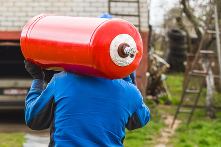 A male industrial worker walks with a gas cylinder to a gas car. Transportation and installation of a propane bottle to residential buildings.の写真素材