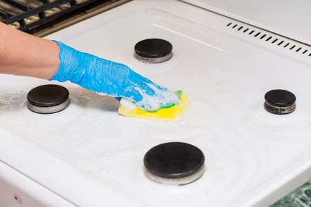 Female hand of a housewife in a household latex glove washes the gas stove with a foaming sponge, close-up.の写真素材