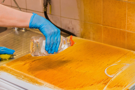Woman's housewife hand in a household glove pours liquid detergent on a dirty coating gas stove on...