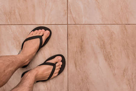 Legs of a man in black flip flops stand on a tiled floor texture background, top view.の写真素材