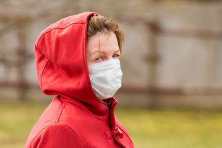 An elderly woman with brown hair and blue eyes in a red coat and hood in a protective safe medical mask, close-up portrait.の写真素材