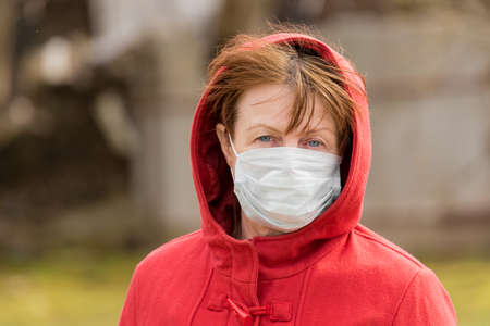 An elderly woman with brown hair and blue eyes in a red coat and hood in a protective safe medical mask, close-up portrait.の写真素材