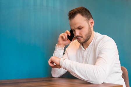 A young European guy businessman in a white shirt, with a beard sits at a table, talks on a mobile phone and looks at the time on a blue background.の写真素材