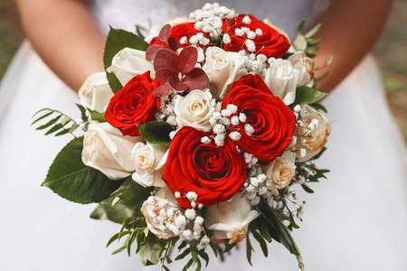 A bride in a wedding dress holds a decorative wedding bouquet of flowers of red and white roses in her hands, close-up.の写真素材