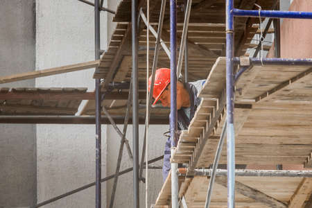 Belarus, Bobruisk - August 21, 2020: An industrial worker in a protective red helmet works safely in the city on scaffolding.のeditorial素材