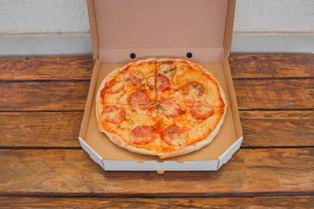 Open cardboard box packaging for freshly made pizza with cheese and salami on a wooden table background.の写真素材