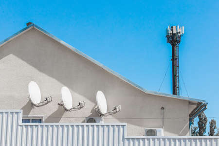 Satellite TV white dishes antenna signal and communications on the wall of a residential building against the background of blue sky and internet tower.の写真素材